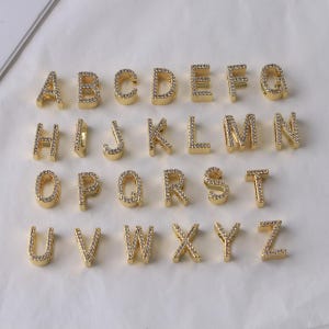 Könnte beinhalten: Goldfarbene Buchstabenanhänger, jeweils mit kleinen, klaren Kristallen besetzt. Die Buchstaben sind in Reihen angeordnet und zeigen das gesamte englische Alphabet von A bis Z. Ideal für die Schmuckherstellung oder personalisierte Bastelarbeiten.