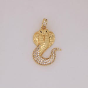 Op de afbeelding: Een goudkleurige cobra-hanger met een gedetailleerd ontwerp. De kop van de cobra is omhoog en het lichaam buigt in een gestileerde S-vorm. De hanger is ingelegd met kleine, heldere edelstenen, wat glans geeft. Ontworpen als sieraad.