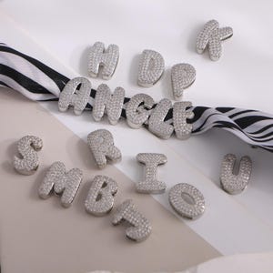 Könnte beinhalten: Silberfarbene Buchstaben im Bubble-Stil, besetzt mit kleinen, funkelnden Steinen. Die Buchstaben sind auf einer weißen Oberfläche mit einem schwarz-weiß gestreiften Band verstreut. Die Buchstaben bilden das Wort ANGLE.