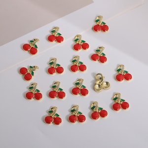 May include: A collection of cherry-shaped charms. Each charm features two red cherries with green leaves and a gold-colored stem. Some stems are adorned with small, clear crystals. The charms are arranged on a white surface.