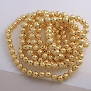 Può includere: Una collezione di perline lucenti di colore dorato, probabilmente per la creazione di gioielli. Le perline sono perfettamente rotonde e di dimensioni uniformi, creando una superficie liscia e riflettente. Le perline sono infilate insieme, formando una collana o un braccialetto.