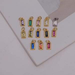 May include: A collection of fourteen rectangular jewelry charms featuring faceted gemstones in various colors including purple, green, pink, clear, blue, black, and orange, each set in a gold-toned metal frame with a small circular jump ring.
