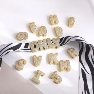 Puede incluir: Letras del alfabeto doradas, estilo burbuja, y la palabra "ONLY" están dispuestas sobre una superficie blanca. Cada letra está incrustada con pequeños cristales transparentes. Una cinta con estampado de cebra en blanco y negro está en el fondo.