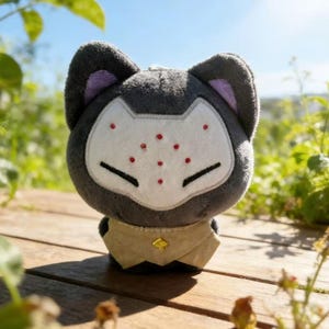 May include: A plush toy cat with a gray body, purple inner ears, and a white face mask with red dots. It has a tan collar with a gold emblem. The toy is sitting on a wooden surface.