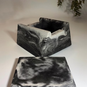 May include: A square, marbled concrete box with a matching lid. The box has a dark gray and white swirling pattern. The lid is rectangular and rests in front of the box. The box has a small notch on the side.