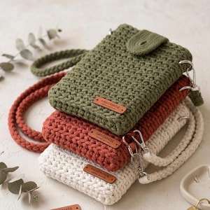 以下が含まれることがあります： オリーブグリーン、ラスト、クリーム色の手作りのかぎ針編み携帯電話バッグが3つ重ねられています。各バッグにはお揃いのストラップと「HANDMADE」と書かれた小さな革製のタグが付いています。バッグは積み重ねられています。