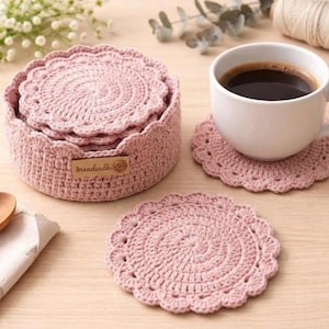 May include: A collection of light pink crochet coasters and a matching storage basket. The coasters have a scalloped edge and are shown with a white coffee cup filled with dark coffee. The basket has a small wooden tag that reads "mandiadie".