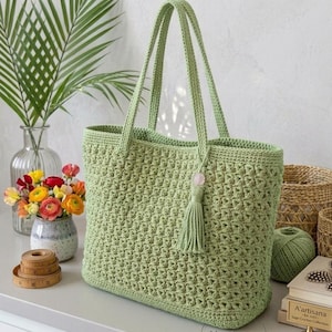 Puede incluir: Bolso tote de ganchillo verde claro con asas largas y una borla decorativa. El bolso tiene un patrón de puntada texturizado y una pequeña cuenta rosa en la borla. El bolso está sobre una superficie blanca.