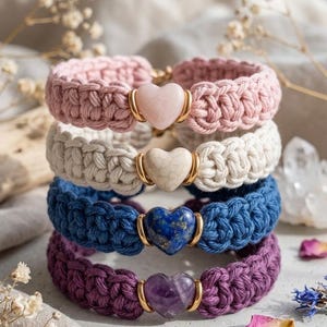 Puede incluir: Cuatro pulseras de macramé hechas a mano en rosa, blanco, azul y morado. Cada pulsera presenta una piedra preciosa en forma de corazón en el centro, enmarcada por metal dorado. Las pulseras están apiladas, mostrando los diferentes colores y texturas.