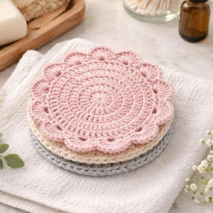 May include: Three handmade crochet coasters in pastel shades. The top coaster is pale pink, the middle is cream, and the bottom is grey. Each coaster features a scalloped edge and a spiral pattern.
