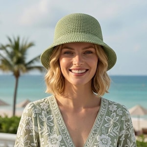 May include: A light green crochet bucket hat with a wide brim. The hat is worn by a person smiling in front of a beach scene. The person is wearing a light green floral patterned top.