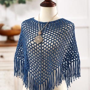 Pode incluir: Um poncho azul de crochê com franjas, exibido em um manequim. O design de trama aberta é combinado com um colar com pingente dourado. O poncho é um acessório elegante.
