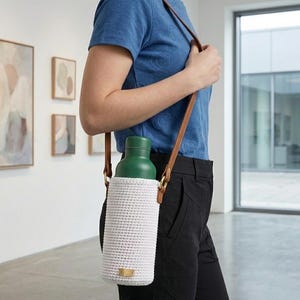 May include: A green water bottle in a white crocheted carrier with a brown leather strap. The carrier has a small gold-coloured tag. The strap is worn over the shoulder. The person is wearing a blue top and black trousers.