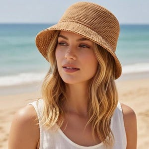 May include: A tan crochet bucket hat with a wide brim. The hat is made of a textured, woven material. The hat is worn by a person with blonde hair, on a beach with the ocean in the background.