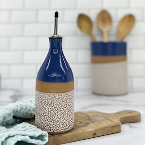 May include: A blue and white ceramic oil and vinegar bottle with a stainless steel pour spout. The bottle has a crackled glaze finish and is sitting on a wooden cutting board.