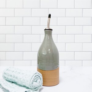 K&ouml;nnte beinhalten: Eine graue Keramik-Oliven&ouml;lflasche mit braunem Keramikfu&szlig; und silbernem Ausgie&szlig;er. Die Flasche steht auf einer wei&szlig;en Arbeitsplatte neben einem hellblauen K&uuml;chentuch.