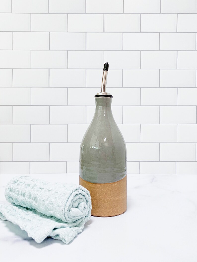 K&ouml;nnte beinhalten: Eine graue Keramik-Oliven&ouml;lflasche mit braunem Keramikfu&szlig; und silbernem Ausgie&szlig;er. Die Flasche steht auf einer wei&szlig;en Arbeitsplatte neben einem hellblauen K&uuml;chentuch.