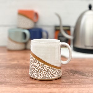 May include: White ceramic mug with a brown speckled glaze on the bottom half. The mug has a rounded handle.