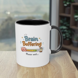 Peut inclure: Mug en céramique blanche avec une anse et un intérieur noirs. Le mug affiche les mots "Brain Buffering" en bleu et orange, ainsi qu'un cerveau de dessin animé et une barre de progression. La phrase "Please wait..." est en dessous.