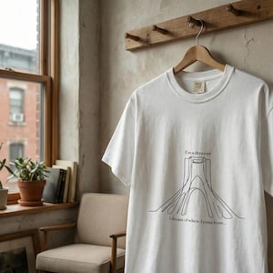 Op de afbeelding: Wit t-shirt met een zwarte lijntekening van een structuur en de tekst "I'm a dreamer" en "I dream of where I come from...". Het shirt hangt aan een houten hanger.
