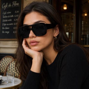 Pode incluir: Óculos de sol pretos de tamanho grande com uma armação espessa de topo plano e um design de ponte dupla proeminente. As lentes são escuras e opacas, proporcionando cobertura total. A armação é feita de material preto sólido com hastes largas.