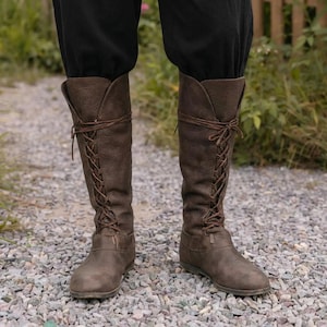 May include: Brown leather knee-high boots with a lace-up design. The boots have a flat sole and a wide opening at the top. The laces are tied in a crisscross pattern up the front of the boots.