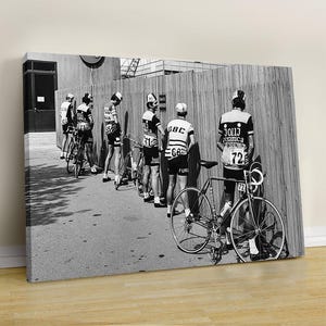 May include: Black and white photograph of a group of cyclists in vintage cycling attire, leaning against a wooden fence. The cyclists are wearing jerseys with numbers and sponsor logos, with their bicycles parked nearby. The image captures a moment of rest or preparation.