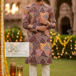 Puede incluir: Un hombre con una túnica estampada de manga larga y pantalones blancos. La túnica presenta un diseño de patchwork en tonos marrones, rosas y amarillos. La imagen está ambientada al aire libre con un fondo borroso de un edificio y vegetación.