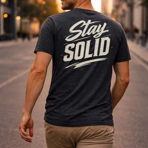 Könnte beinhalten: Dunkelgraues T-Shirt mit dem weißen Schriftzug "Stay Solid" in stilisiertem Text. Das Shirt hat kurze Ärmel und wird von einer Person getragen, die eine Straße entlanggeht. Die Person trägt eine khakifarbene Hose.