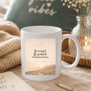 Könnte beinhalten: Weiße Keramiktasse mit Landschaftsdesign und dem Text "Be present. Be grateful. Create your future." Die Tasse hat einen gebogenen Griff und steht auf einem Tisch mit Stift und Notizblock.