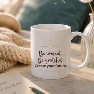 Könnte beinhalten: Weiße Keramik-Tasse mit dem Text "Be present. Be grateful. Create your future." in brauner Schrift. Die Tasse hat ein goldenes Sonnendesign und florale Akzente. Ein Stift und ein Notizblock befinden sich im Vordergrund.