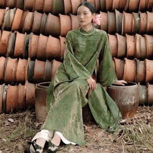 Peut inclure: Une tunique vert olive longue jusqu'au sol &agrave; manches longues avec un motif textur&eacute; subtil, port&eacute;e sur un pantalon blanc. Le v&ecirc;tement pr&eacute;sente une encolure haute, une fente lat&eacute;rale, un collier argent&eacute; simple et des chaussures plates &agrave; motifs.
