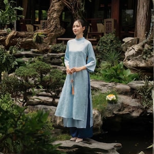 May include: A light blue long-sleeved tunic with a subtle tonal floral pattern and high side slits, paired with dark blue wide-leg trousers. The tunic features a high collar and a small decorative tassel hanging from the center front area.