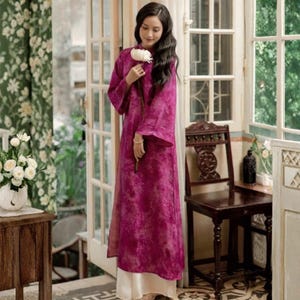 Peut inclure: Une robe magenta &agrave; manches longues avec un motif ton sur ton subtil, dot&eacute;e d'un col montant et d'une silhouette ample et fluide descendant jusqu'aux chevilles, port&eacute;e sur une jupe blanche unie, avec une fleur blanche tenue pr&egrave;s du buste.