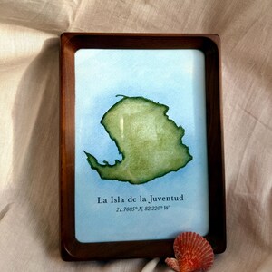 Może przedstawiać: Oprawiona akwarela przedstawiająca mapę La Isla de la Juventud, Kuba, w odcieniach zieleni i błękitu. Ciemna drewniana rama otacza dzieło, z nazwą wyspy i współrzędnymi poniżej. Mała, pomarańczowa muszla spoczywa w pobliżu ramy.