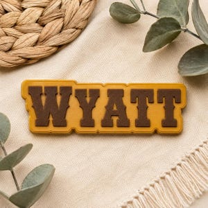 May include: A rectangular name sign featuring the name WYATT in dark brown, block-style capital letters. The letters are set against a mustard yellow background with a scalloped, raised border that follows the contour of the text.
