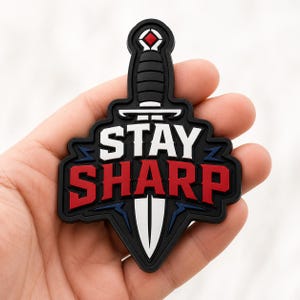 May include: A black, white, and red rubberized patch shaped like a dagger. The text STAY SHARP is printed in bold white and red lettering across the center. The design features a stylized blade and handle with a small red diamond accent at the top.
