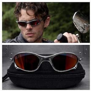 May include: Silver-framed sunglasses with red lenses, presented on a black case. The sunglasses have a sleek, modern design. The image also shows a person wearing the sunglasses.