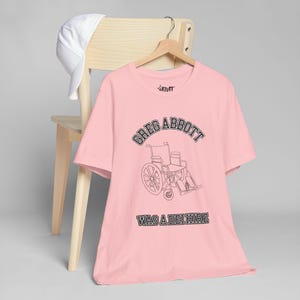Puede incluir: Una camiseta rosa claro con el texto "GREG ABBOTT" encima de un dibujo de una silla de ruedas y las palabras "WAS A DEI HIRE" debajo. La camiseta está colgada en una percha de madera sobre una silla de madera, con una gorra blanca en la silla.