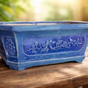 Peut inclure: Un pot de fleurs rectangulaire en céramique bleue avec une finition vieillie. Le pot présente des motifs floraux et d'oiseaux en relief sur les côtés. Il repose sur une surface en bois, idéal pour exposer des plantes. Le pot mesure environ 30 cm de long.
