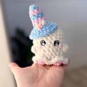 May include: A small, cream-colored crocheted ghost plushie featuring a light blue pointed hat decorated with pink and yellow flower appliques. The ghost has large black plastic safety eyes with white highlights and small pink embroidered cheeks and feet.