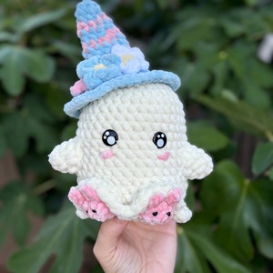 May include: A white crocheted ghost wearing a blue and pink crocheted witch hat with a white flower accent. The ghost has black eyes and pink cheeks.