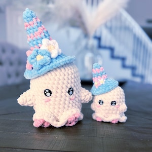 May include: Two cream-colored crochet ghost figures with large black eyes and pink cheeks. Each ghost wears a tall, pointed hat in a variegated blue and pink yarn, decorated with small white and blue crocheted flowers at the base of the hat.