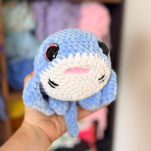May include: A handmade blue and white plush jellyfish toy with black eyes, pink accents, and a smiling mouth. The jellyfish has a soft, textured appearance and is held in a person's hand.