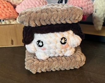 May include: A handmade crochet s'more plush toy. The s'more features a white marshmallow with black eyes and pink cheeks, sandwiched between two tan graham crackers and a dark brown chocolate layer. Other crochet plushies are in the background.