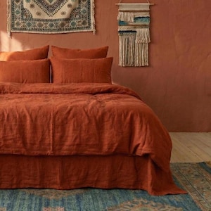 以下が含まれることがあります： 錆色のリネン寝具セット。掛け布団カバー、枕カバー、ベッドスカートが含まれています。寝具は、テラコッタ色のテクスチャード加工された壁を背景にベッドに配置されています。装飾的な壁掛けがボヘミアンな雰囲気を醸し出しています。