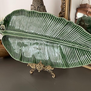 May include: A glossy, sage green ceramic serving platter shaped like a large, elongated tropical leaf. The platter features a detailed, raised vein pattern across its surface and a curled stem at one end, displayed on an ornate gold metal stand.