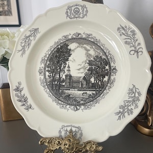 May include: A cream-colored ceramic plate with a scalloped edge, featuring a central black and white transfer print of a church landscape. The rim is decorated with botanical illustrations and small crests at the top and bottom positions.