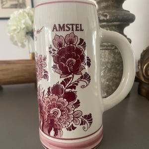 May include: A white ceramic beer stein featuring a dark red floral pattern and the word AMSTEL printed in dark red near the top. The stein has a cylindrical body, a sturdy side handle, and horizontal red bands around the base.
