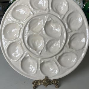 May include: A white ceramic oyster serving platter with 12 individual shell-shaped compartments. The platter is round and sits on a decorative gold-colored metal stand. The design is elegant and suitable for serving seafood.